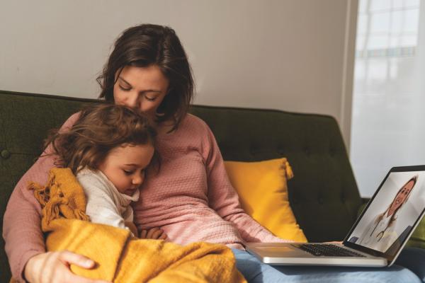 Madre che conforta la sua giovane figlia, sedute su un divano con un laptop che mostra un medico sullo schermo.